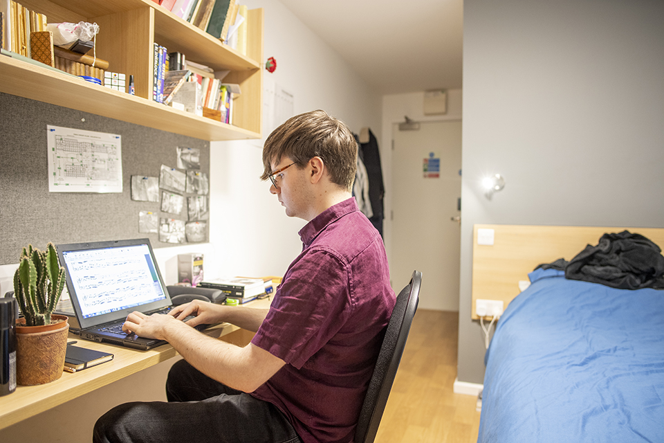 A male student looking at his laptop, sitting in a bedroom.